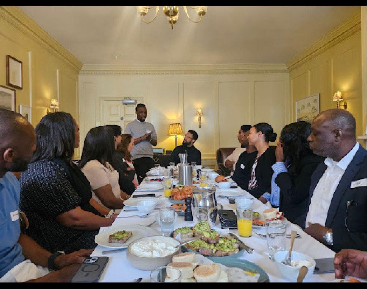Emperor Roy speaking at a breakfast event, standing at the head of a table